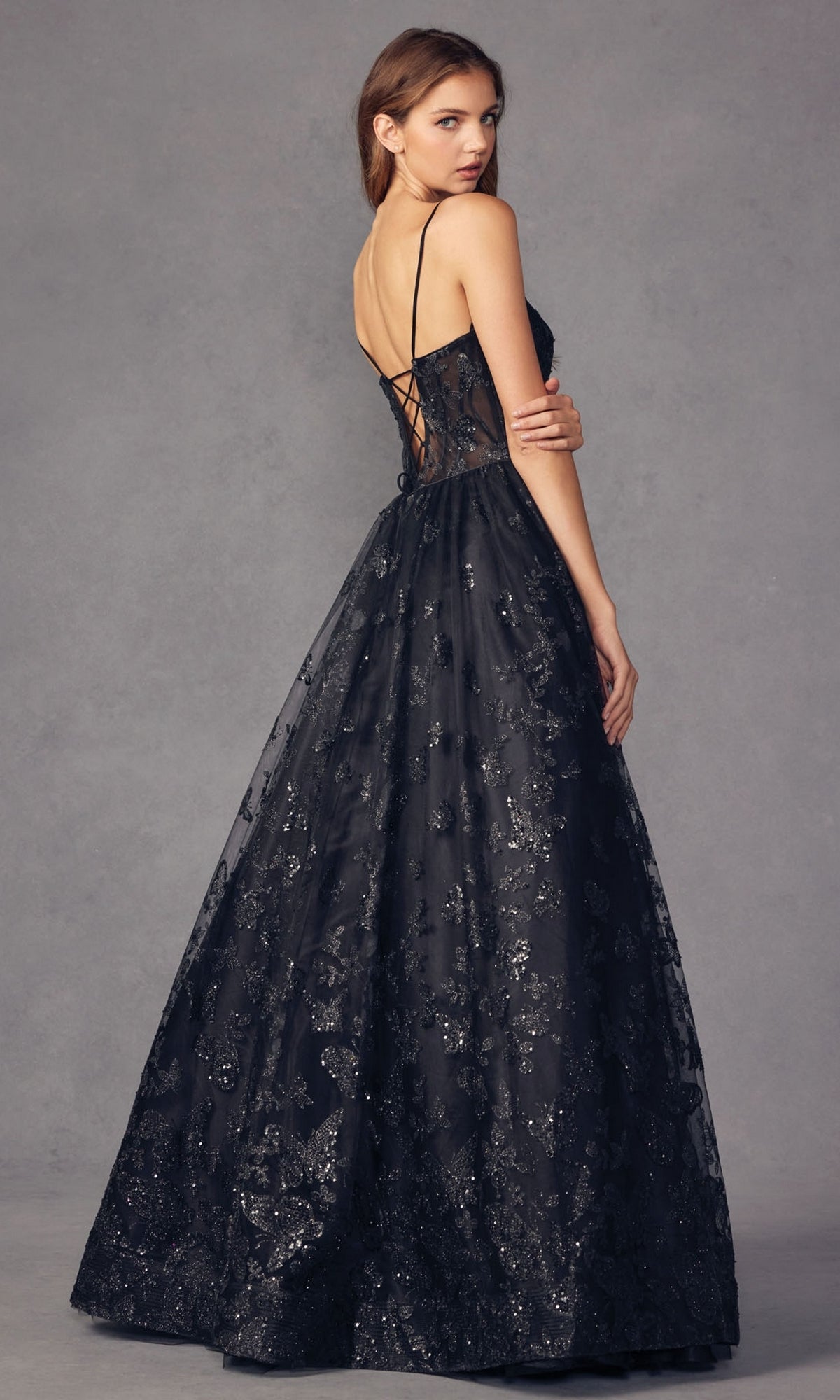 A woman in the Sheer-Corset Glitter-Print Long Prom Dress 2413, featuring a lace-up back and subtle glitter patterns, stands against a gray background, looking over her shoulder.